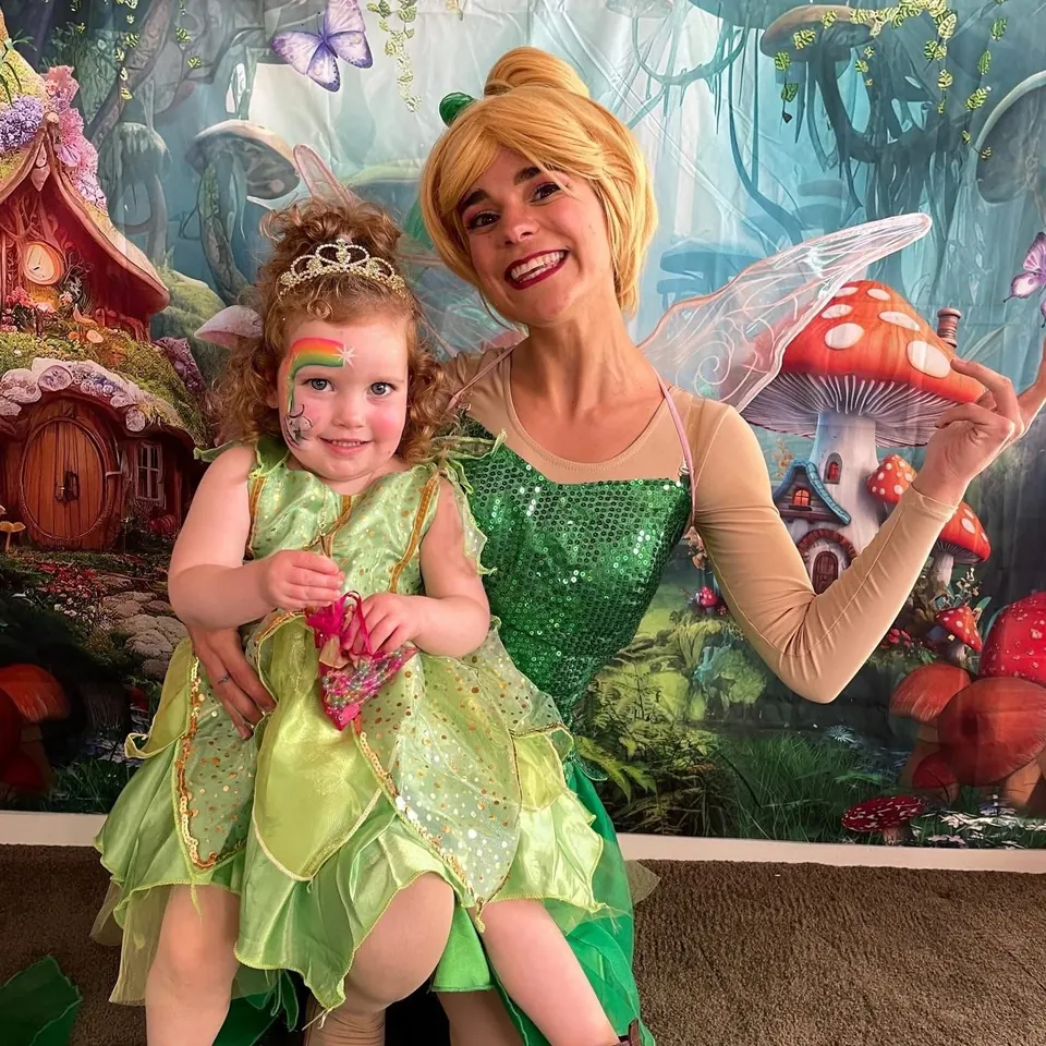 Tinkerbell fairy party entertainer in green sequin dress with wings cuddling toddler in matching Tinkerbell costume and rainbow face paint at a Melbourne fairy themed party