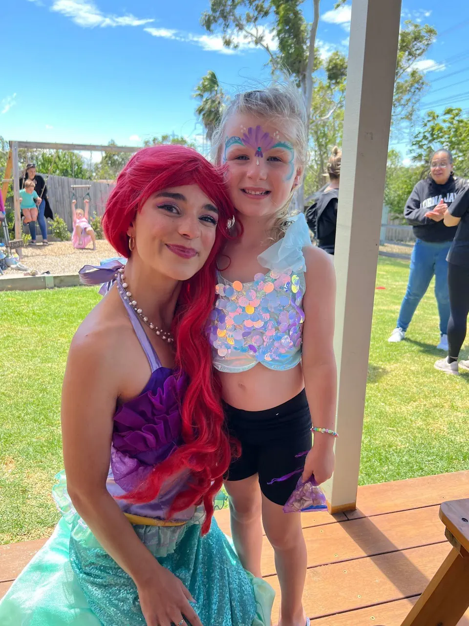 Little Mermaid Ariel party entertainer with red hair and sparkly tail posing with smiling girl wearing mermaid face paint at a Melbourne backyard birthday