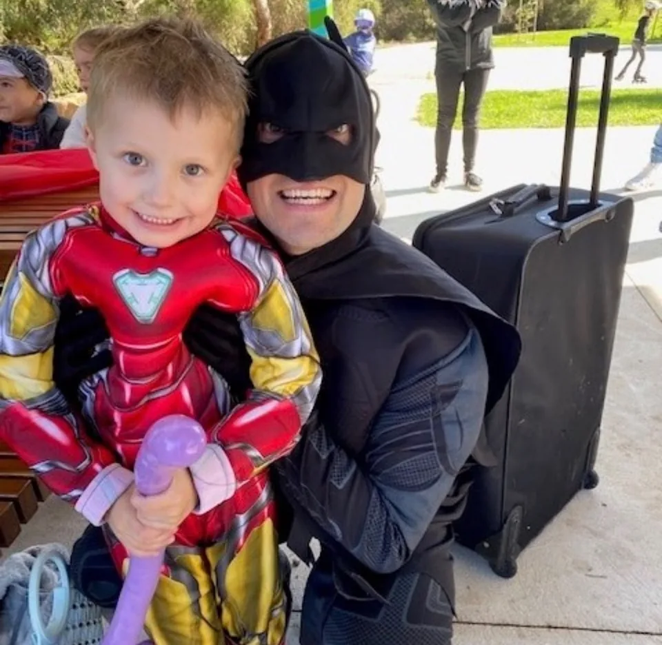 Batman party entertainer in black armoured suit and cowl smiling cheek-to-cheek with young boy in Iron Man costume holding a purple balloon at a Melbourne park superhero birthday party