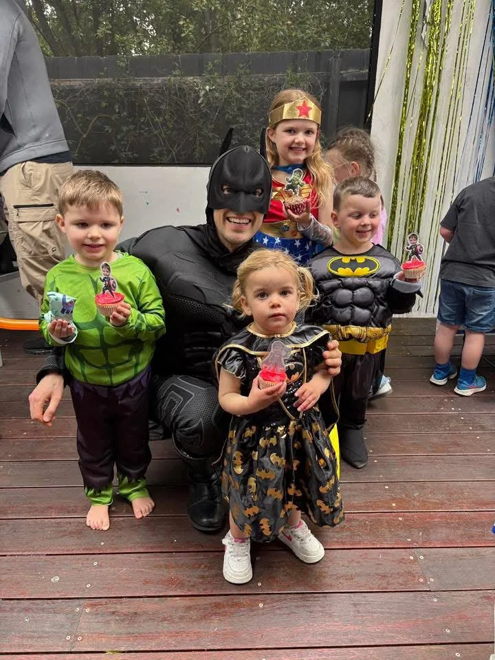 Batman party entertainer in black muscle suit and cowl crouching with a group of young kids in Hulk, Wonder Woman, Batman and Batgirl costumes holding superhero cupcakes at a Melbourne backyard superhero birthday party