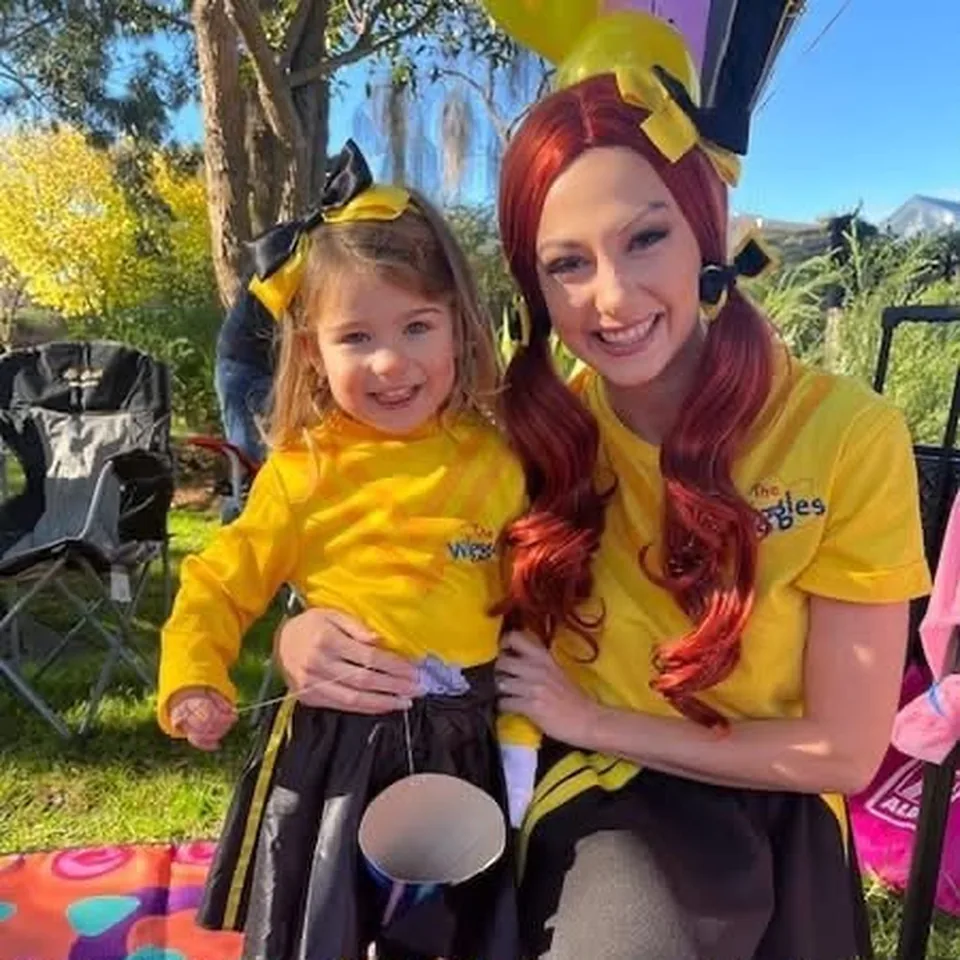 Emma Wiggle character entertainer with red hair and yellow bow smiling with toddler in matching yellow Wiggles top and bow at an outdoor Melbourne Wiggles themed birthday party