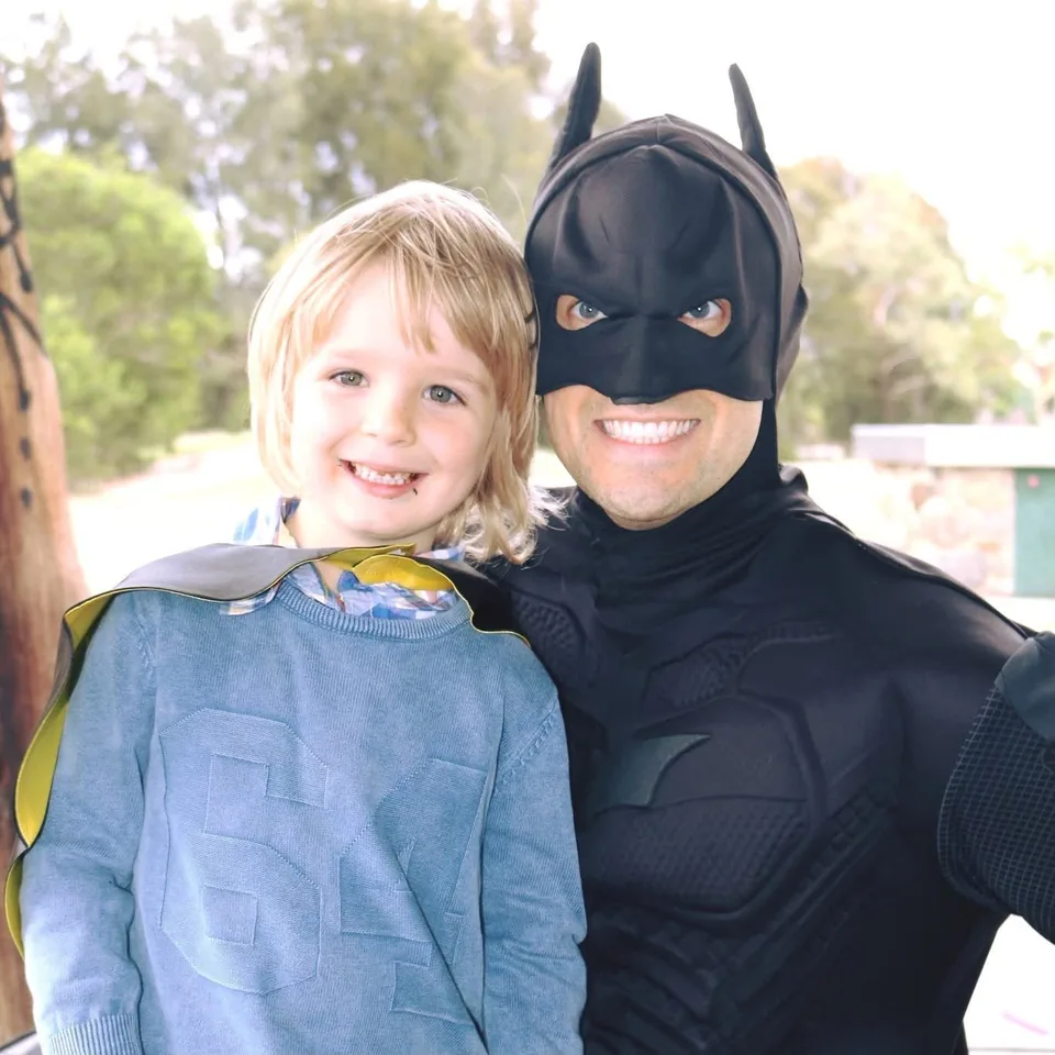 Batman party entertainer in black muscle suit and cowl posing with smiling blonde boy in a Batman cape at a Melbourne park superhero birthday party