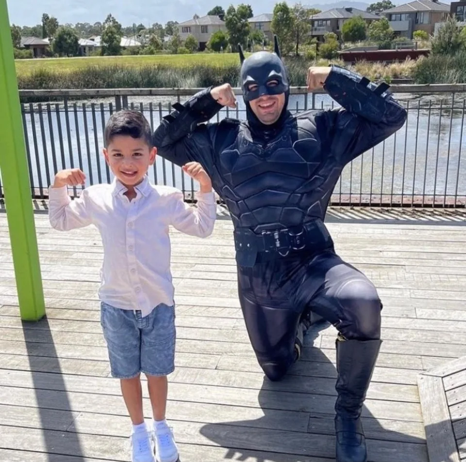 Batman party entertainer in full black muscle suit and cowl flexing muscles beside smiling young boy in a white shirt at a Melbourne lakeside outdoor superhero birthday party