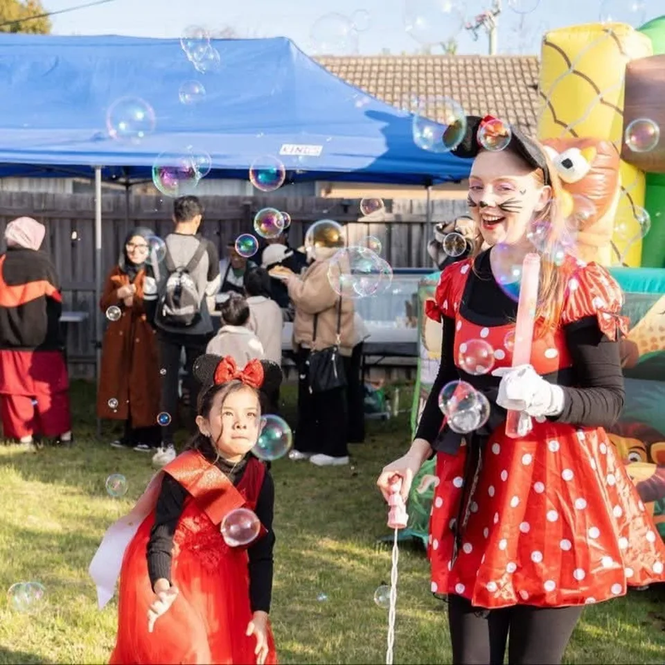 Minnie Mouse party entertainer in red polka dot dress and cat face paint blowing bubbles for young girl in red dress and Minnie ears at a Melbourne backyard Minnie Mouse birthday party