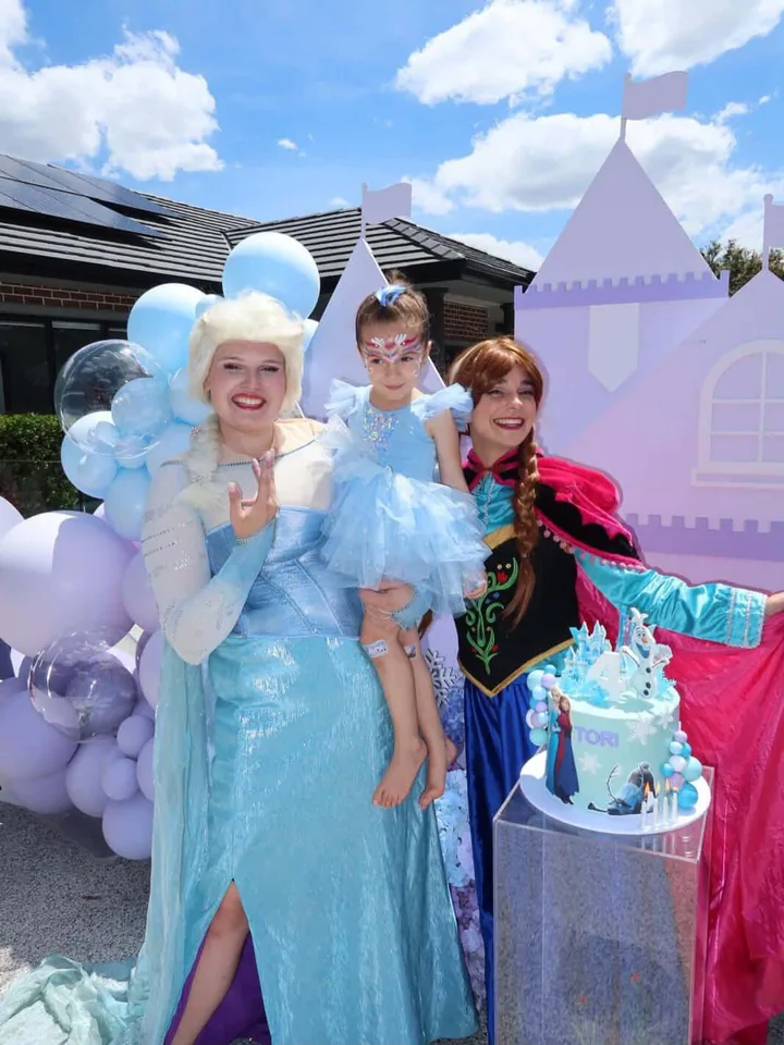 Frozen Elsa and Anna party entertainers holding birthday girl in a blue ruffled dress with snowflake face paint in front of a purple castle backdrop, blue balloon arch and Tori's 4th birthday Frozen cake at a Melbourne Frozen themed birthday party