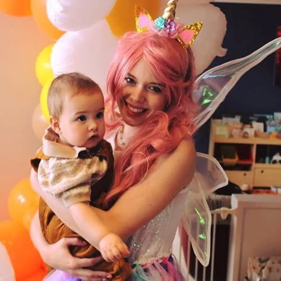 Unicorn fairy party entertainer with pink hair, gold unicorn horn and iridescent wings cuddling a baby in a brown outfit at a Melbourne first birthday unicorn themed party