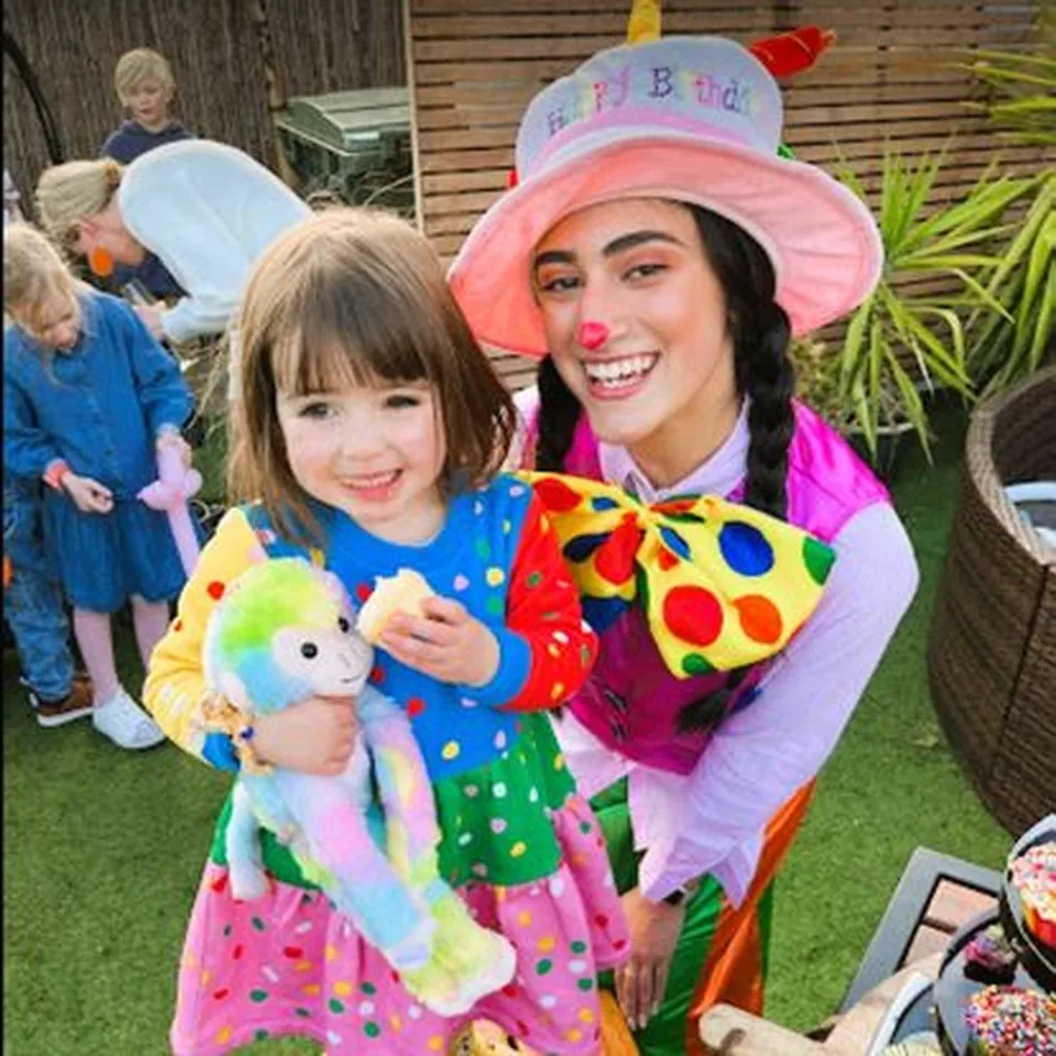 Bubbles the Clown party entertainer in pink birthday hat smiling with young girl holding rainbow plush monkey at a Melbourne clown themed kids birthday party