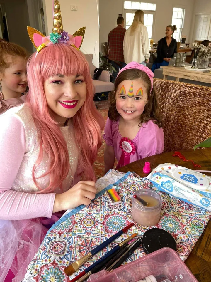 Unicorn face painter party entertainer with pink hair and gold unicorn horn headband smiling beside young birthday girl wearing rainbow unicorn face paint at a Melbourne unicorn themed birthday party