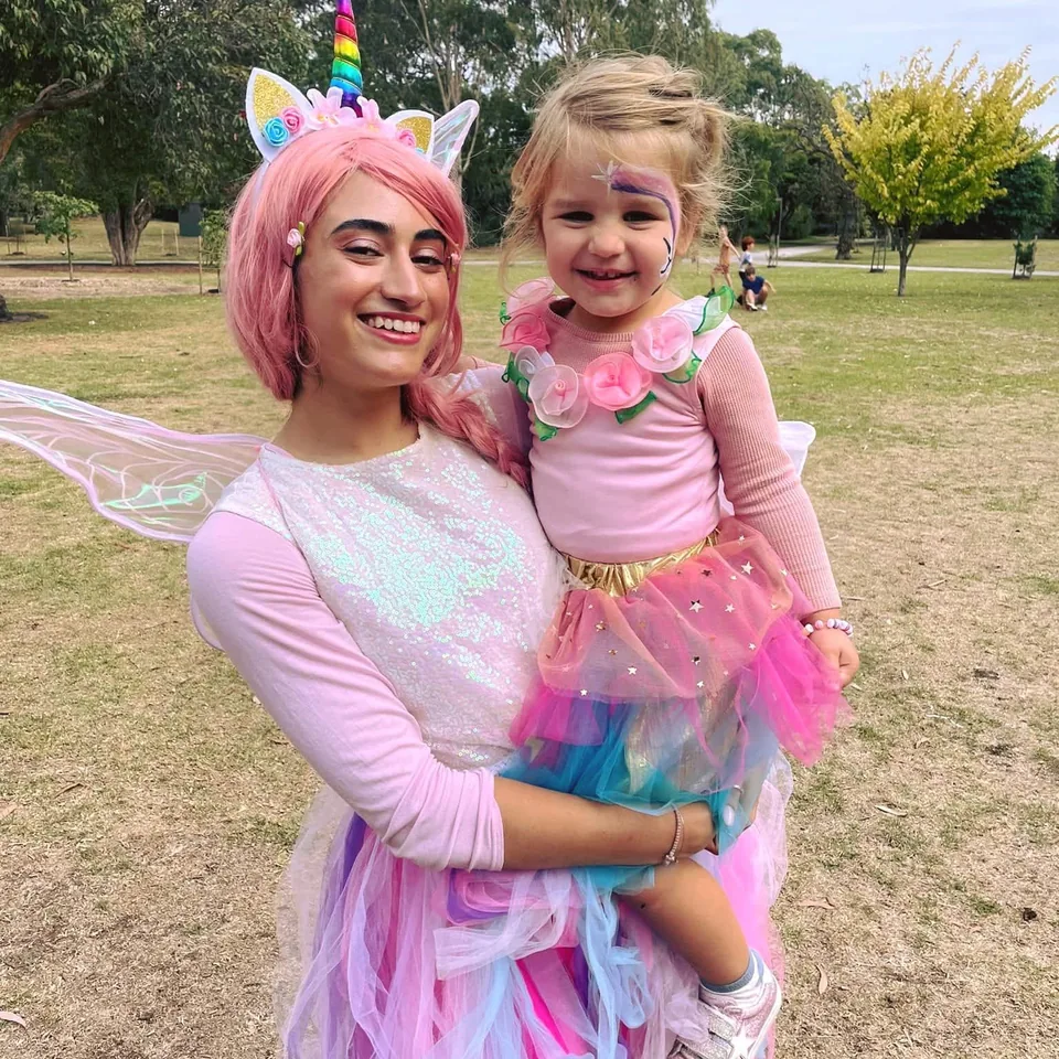 Unicorn fairy party entertainer with pink hair, rainbow horn and shimmering wings cuddling toddler in pink rose tutu and unicorn face paint at a Melbourne park birthday party