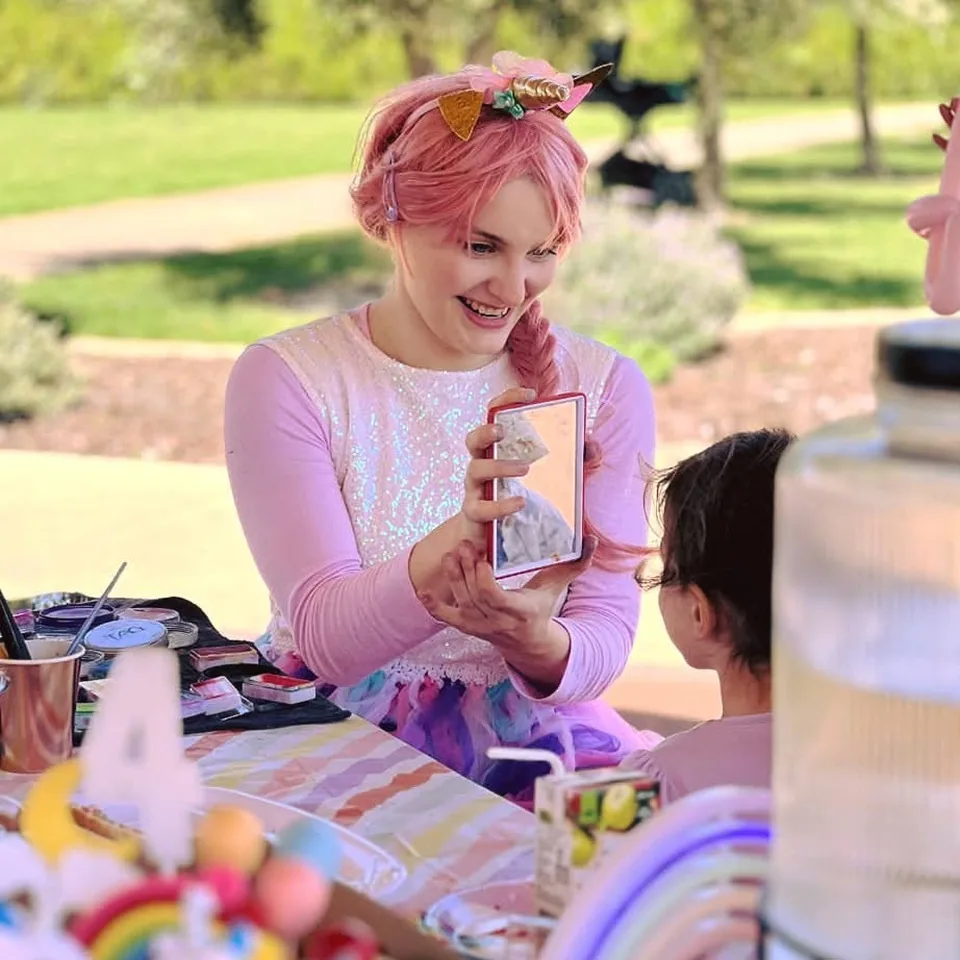 Unicorn fairy face painter party entertainer with pink hair, gold unicorn horn and sparkly dress smiling as she holds a mirror up for a child after face painting at a Melbourne unicorn themed park birthday party