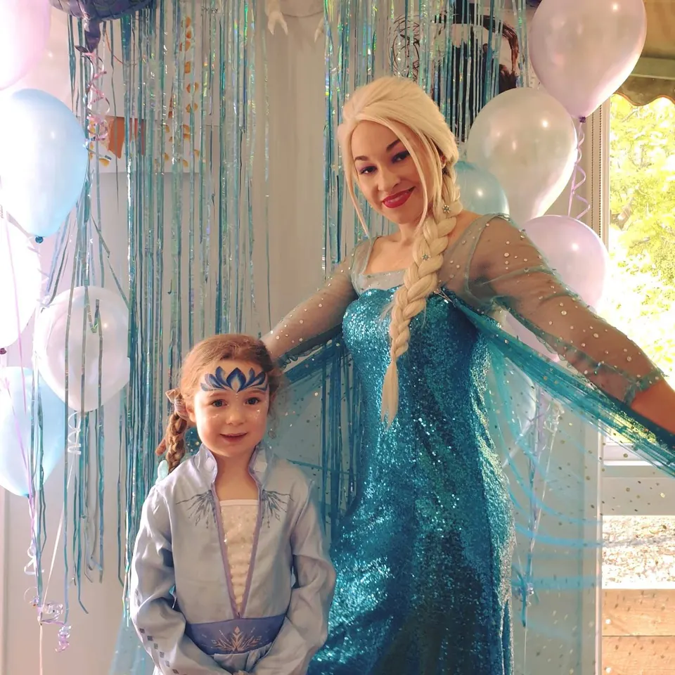 Frozen Elsa party entertainer in sparkling blue gown with young girl in Elsa costume and snowflake face paint in front of icicle backdrop and balloons in Melbourne