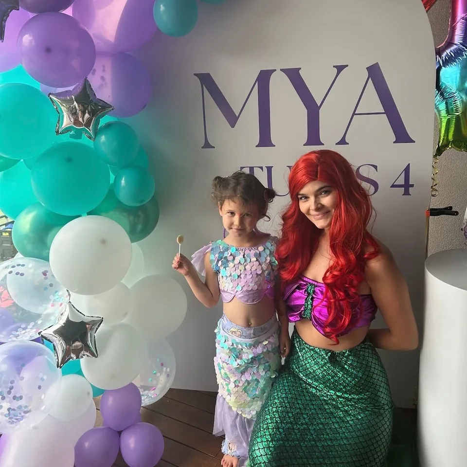 Little Mermaid Ariel party entertainer with red wig and green sequin tail posing with young girl in sequin mermaid costume beside teal and purple balloon arch at Mya's 4th Melbourne mermaid birthday party