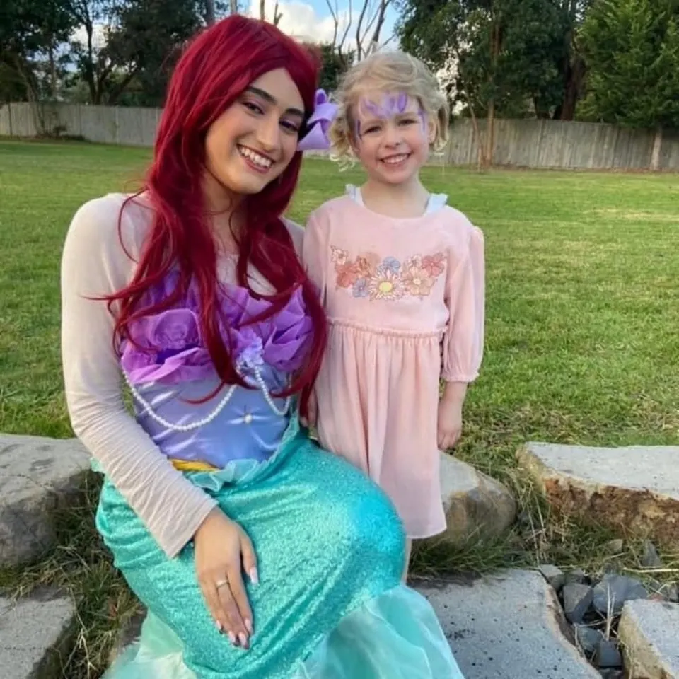 Little Mermaid Ariel party entertainer with red wig and teal sequin tail sitting with smiling girl in pink dress at a Melbourne backyard kids birthday party