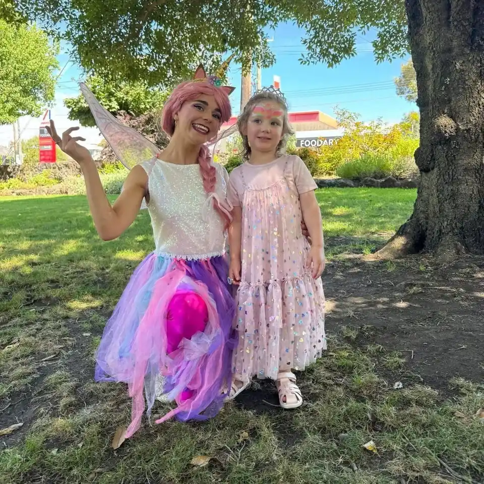 Unicorn fairy party entertainer with pink hair, gold horn and fairy wings posing in a sunny park with young girl in tiara, sparkly pink dress and rainbow unicorn face paint at a Melbourne outdoor unicorn fairy birthday party