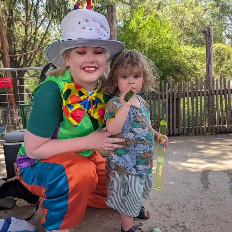 Birthday clown party entertainer in colourful costume and cake hat blowing bubbles with toddler in dinosaur shirt at Melbourne Zoo party