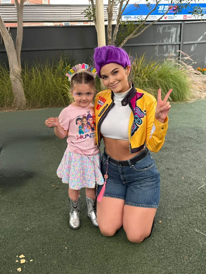 K-Pop Demon Hunters Rumi party entertainer in purple wig and yellow Huntr/x jacket kneeling with toddler birthday girl in a Huntr/x T-shirt at a Melbourne park birthday party