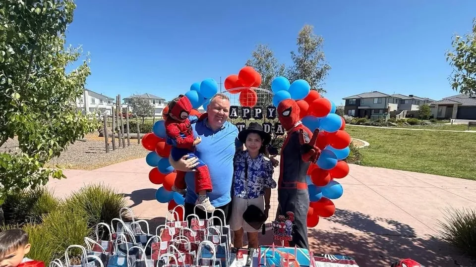 Spider-Man party entertainer posing with family and toddler in Spider-Man costume in front of red and blue balloon arch at a Melbourne park superhero birthday party