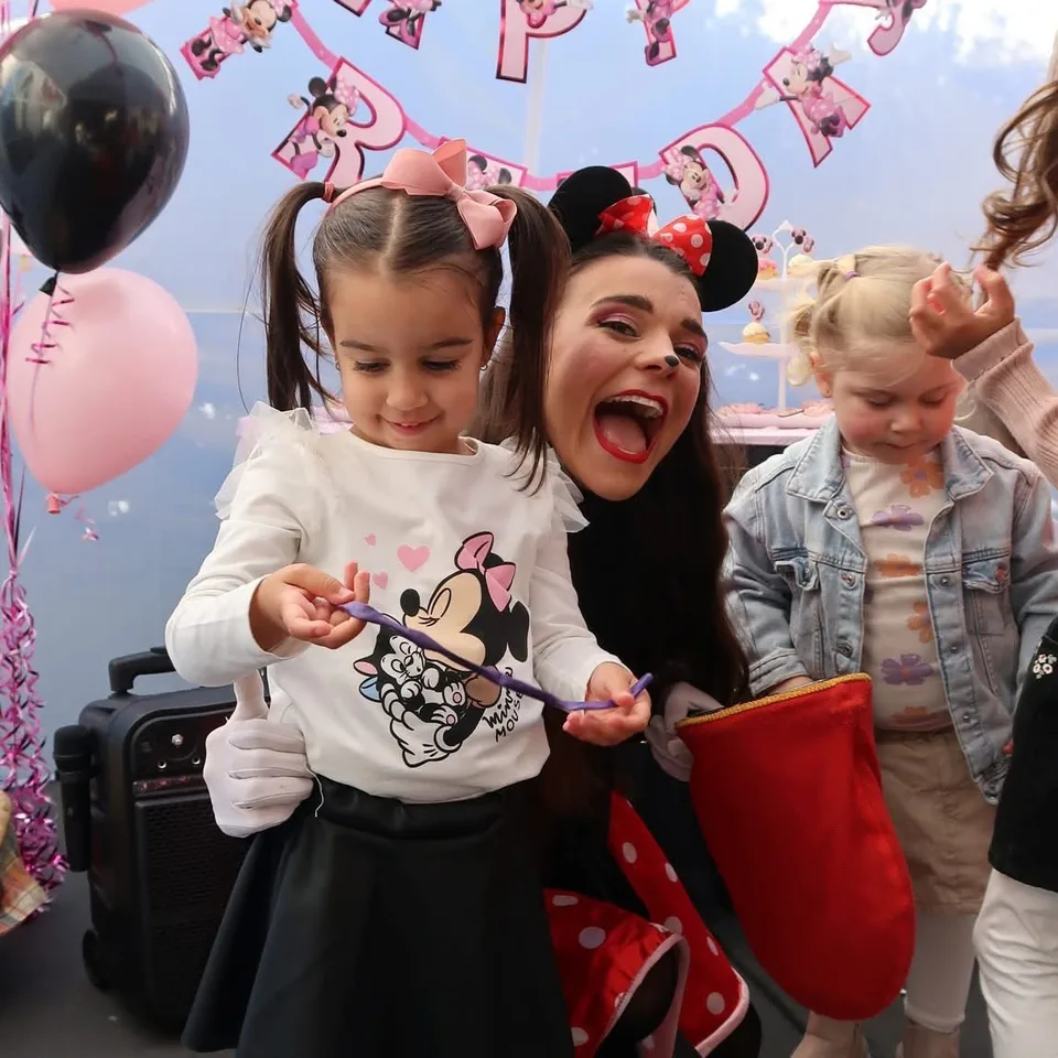 Minnie Mouse party entertainer with mouse ears and red bow making purple balloon animal with smiling girl in pigtails at a Melbourne Minnie Mouse themed birthday party