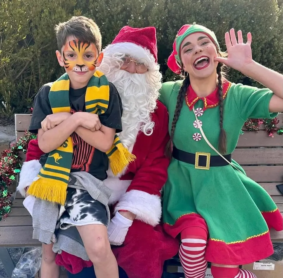 Santa Claus and Christmas elf party entertainers posing with young boy in tiger face paint on a festive bench at a Melbourne Christmas event