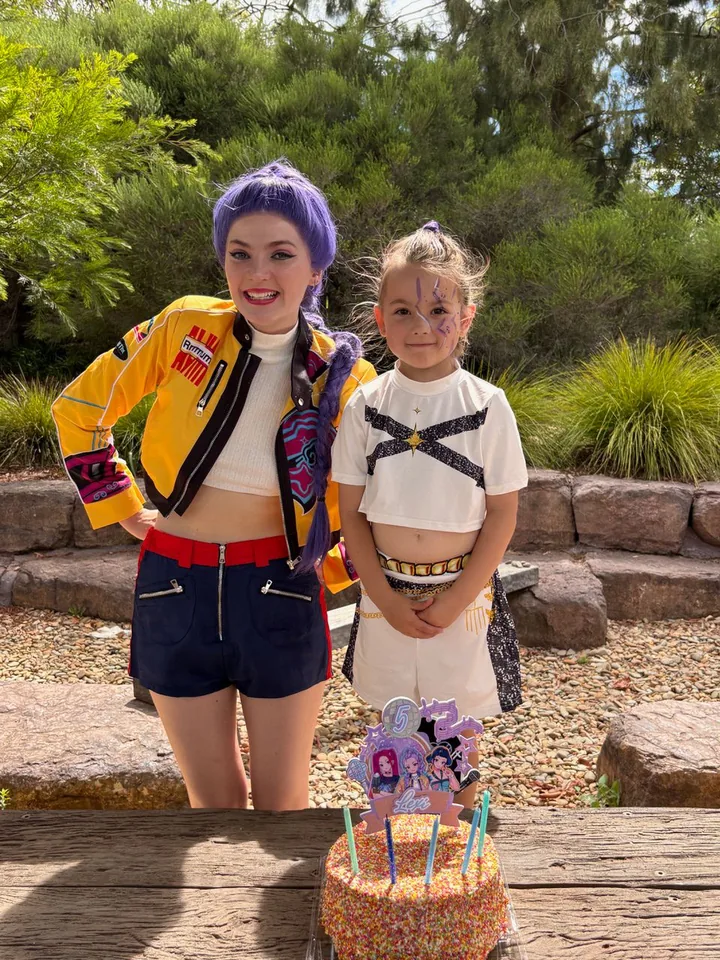 K-Pop Demon Hunters Rumi party entertainer in purple wig and yellow Huntr/x jacket posing with young girl in Huntr/x costume beside sprinkle birthday cake at a Melbourne garden 5th birthday party