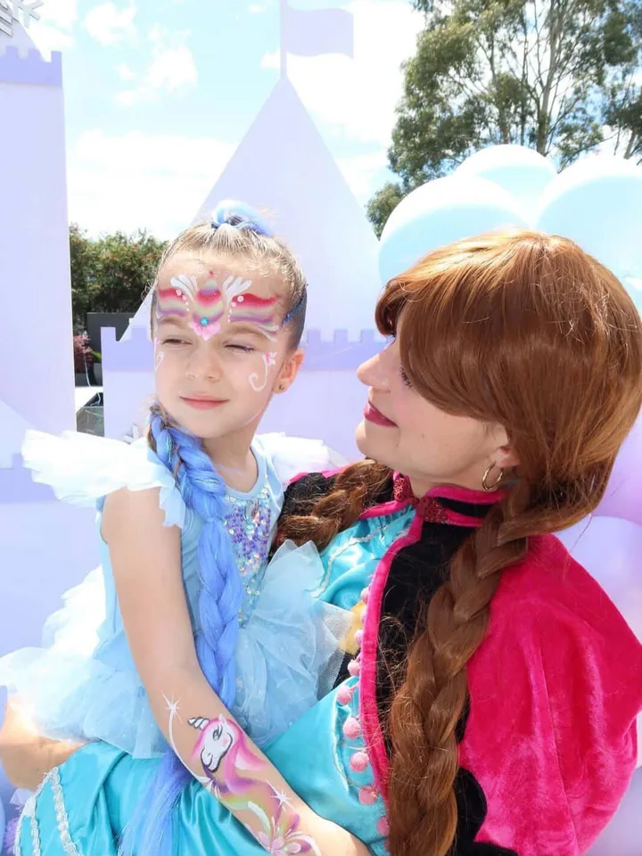 Frozen Anna party entertainer with red braid and pink cape holding young girl in blue sparkly dress with rainbow snowflake face paint and a unicorn arm paint at a Melbourne Frozen unicorn themed birthday party