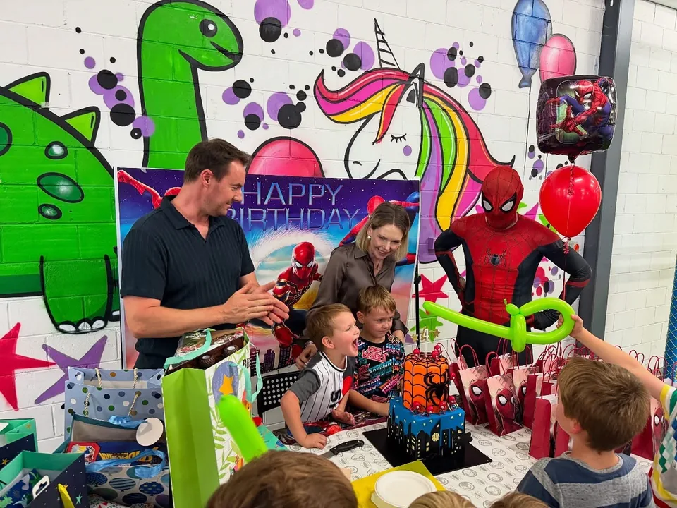 Spider-Man party entertainer making green balloon animals beside birthday boy and Spider-Man cake at a Melbourne indoor superhero birthday party