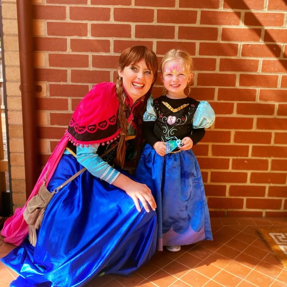 Frozen Anna party entertainer with red braid and pink cape kneeling beside young girl in matching Anna costume with rainbow princess face paint at a Melbourne Frozen birthday party