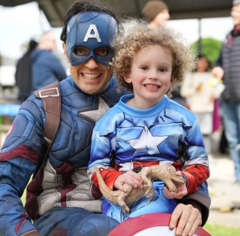 Captain America party entertainer in full muscle suit and mask cuddling a smiling curly-haired boy in Captain America costume holding a bearded dragon lizard at a Melbourne outdoor superhero birthday party