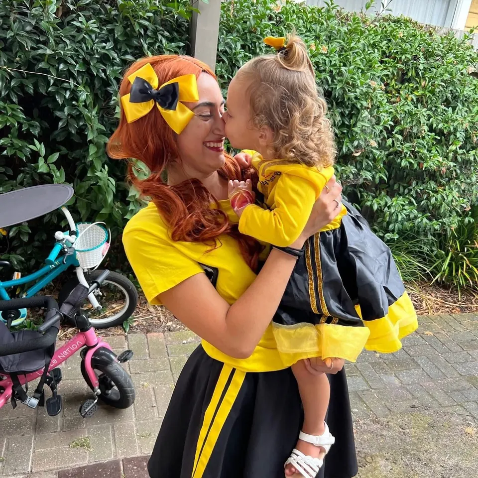 Emma Wiggle character entertainer in yellow bow kissing toddler dressed as Emma at a Wiggles-themed first birthday party in Melbourne