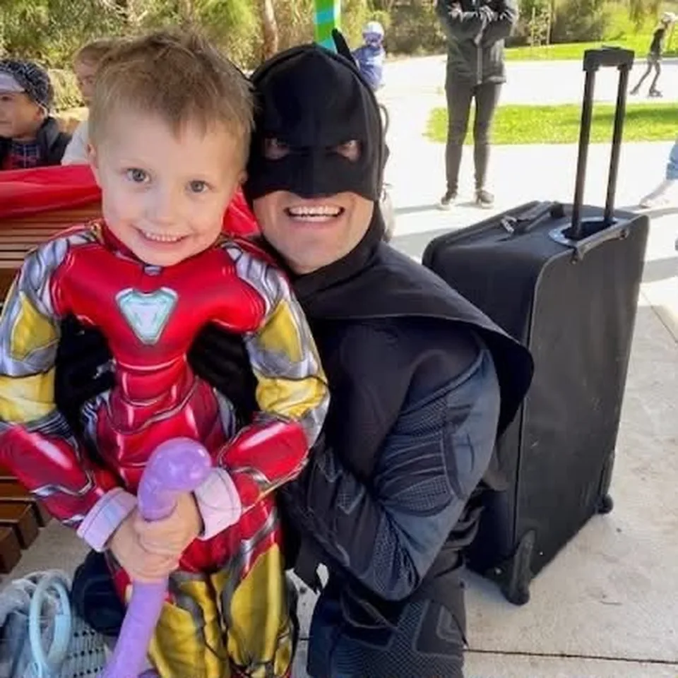 Batman party entertainer in black muscle suit cuddling smiling young boy in Iron Man costume holding a purple balloon animal at a Melbourne outdoor superhero birthday party