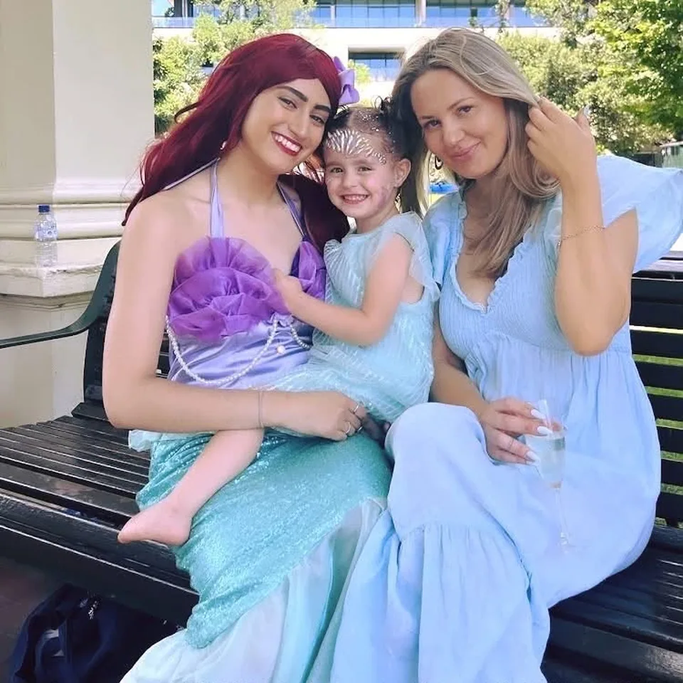 Little Mermaid Ariel party entertainer in purple and teal sequin tail sitting on a bench cuddling young girl in blue dress and pearl tiara with her mum at a Melbourne mermaid birthday party