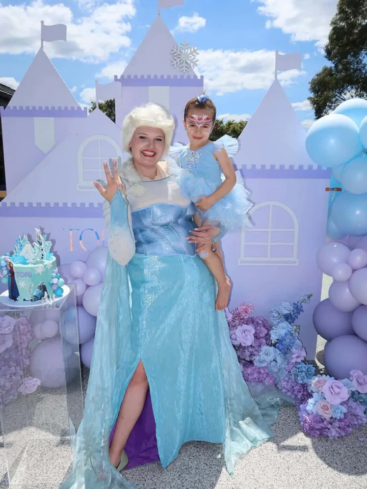 Frozen Elsa party entertainer in sparkling blue gown holding birthday girl in a blue ruffled dress with snowflake face paint in front of a purple Tori castle backdrop and Frozen 4th birthday cake at a Melbourne Frozen themed birthday party