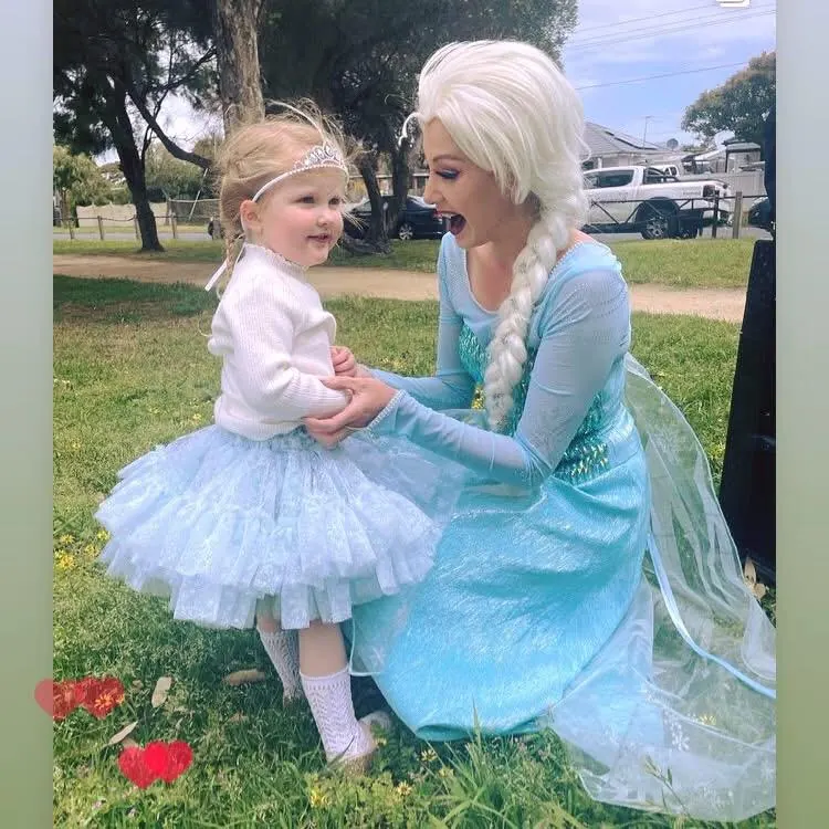 Frozen Elsa party entertainer in blue ice dress crouching with toddler in tiara and tutu on grass at a Melbourne outdoor princess birthday party