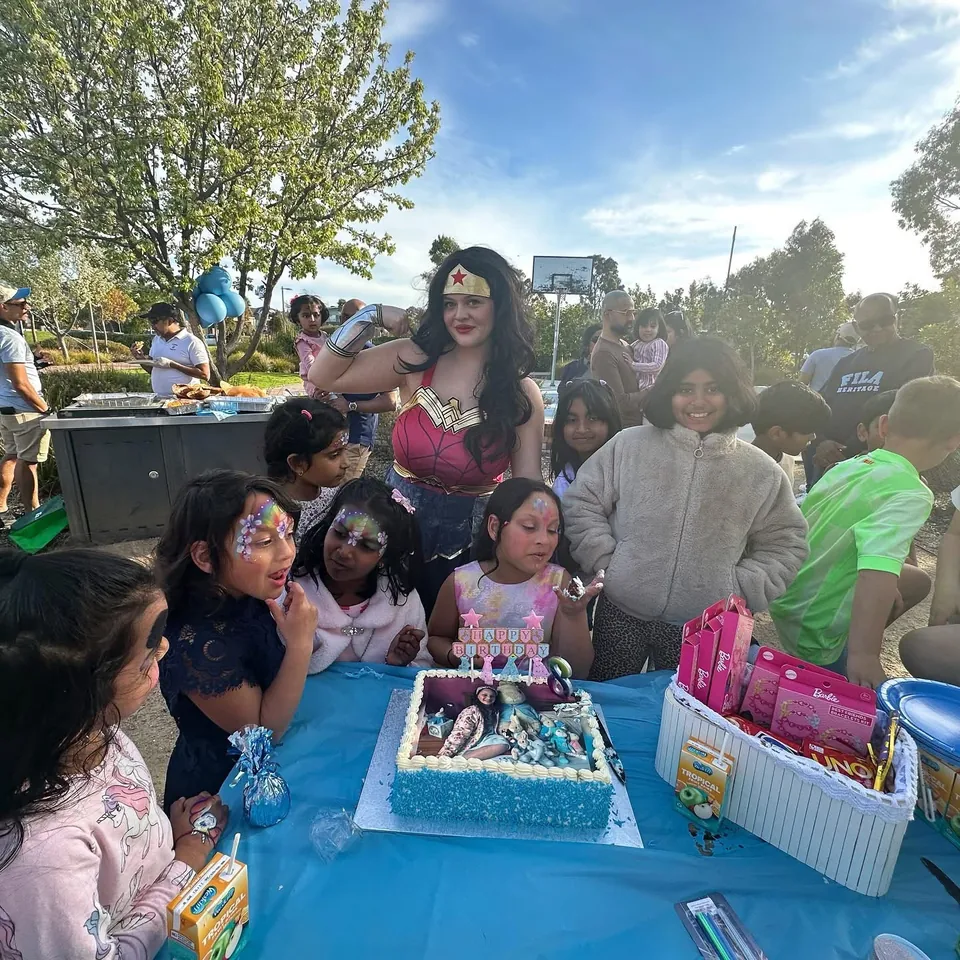 Wonder Woman superhero party entertainer flexing with group of kids in rainbow butterfly face paint around a Frozen birthday cake at a Melbourne park superhero birthday party