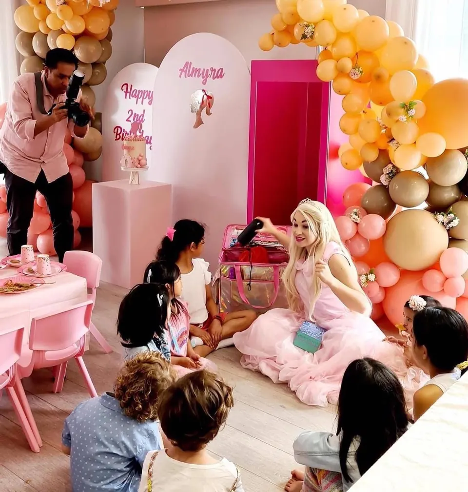 Barbie princess party entertainer in pink tulle dress reading to a group of children at Almyra's 2nd birthday with pink and peach balloon arch