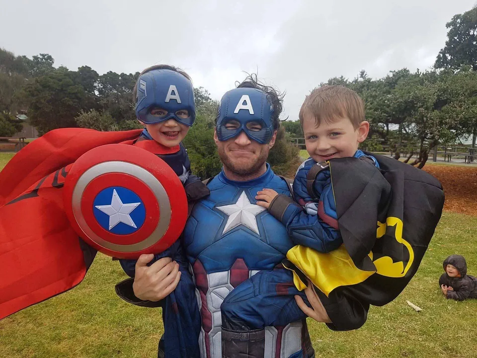 Captain America party entertainer in full muscle suit holding two young boys, one in a Captain America mask and cape and one in a Batman cape, at a Melbourne park superhero birthday party