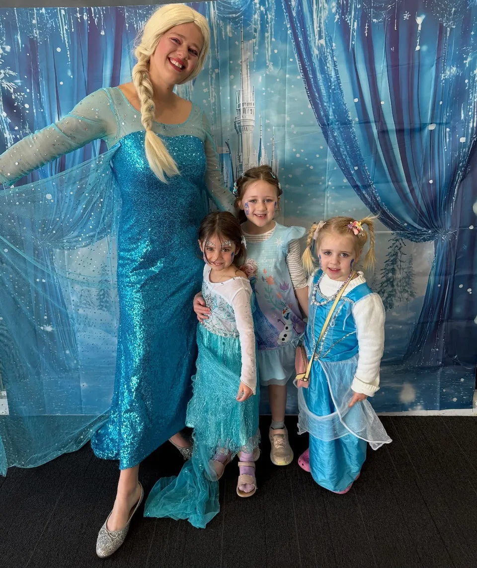 Frozen Elsa party entertainer in sparkling blue gown posing with three young girls in Elsa and princess costumes and snowflake face paint in front of a Frozen ice castle backdrop at a Melbourne princess birthday party