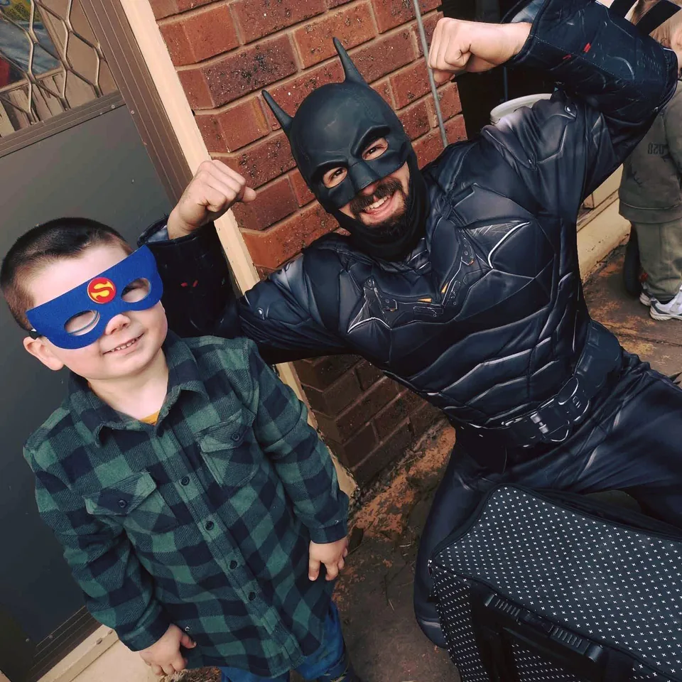 Batman party entertainer in black armoured suit flexing muscles beside smiling young boy in Superman felt mask at a Melbourne front-door superhero birthday party