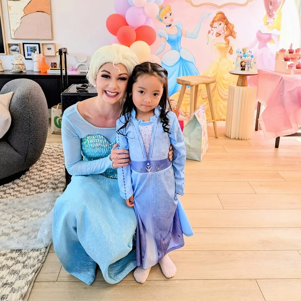 Frozen Elsa party entertainer hugging young girl in Elsa costume in front of Disney princess backdrop and balloon garland at a Melbourne princess party