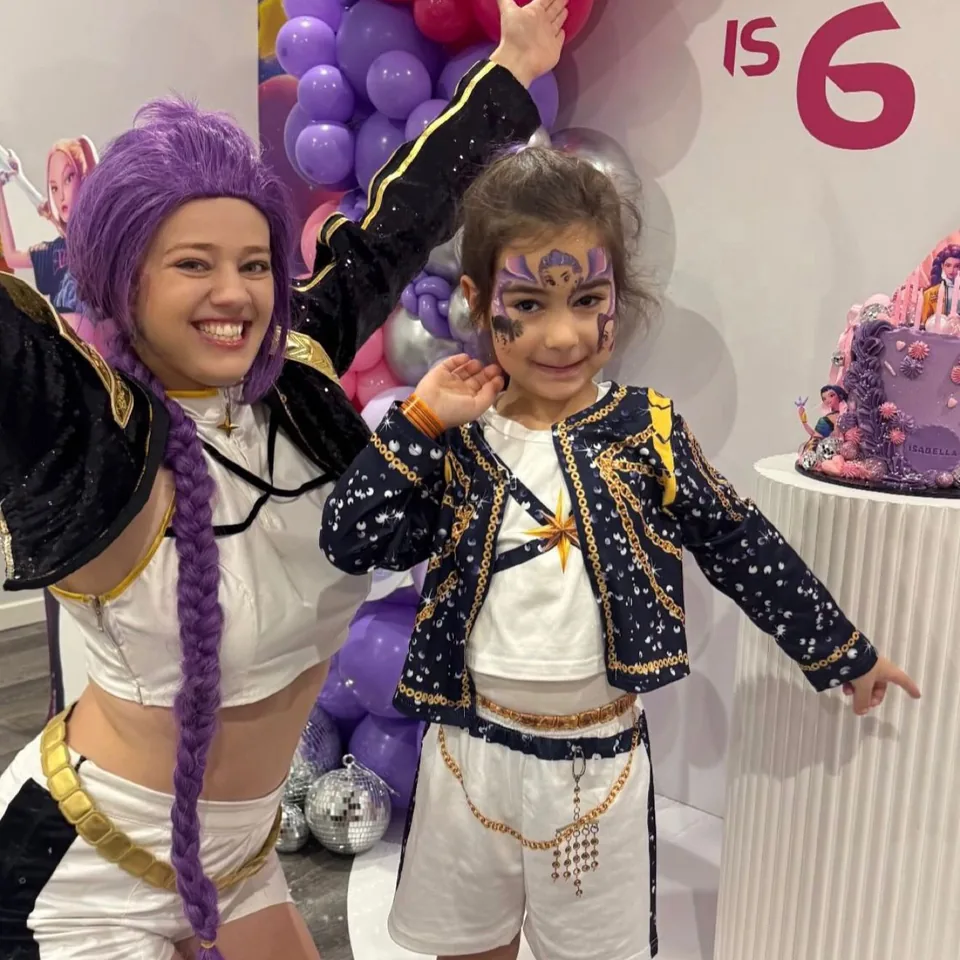 K-Pop Demon Hunters Mira party entertainer with long purple braid dancing with young girl in Huntr/x costume in front of purple balloon arch and birthday cake at a Melbourne 6th birthday party