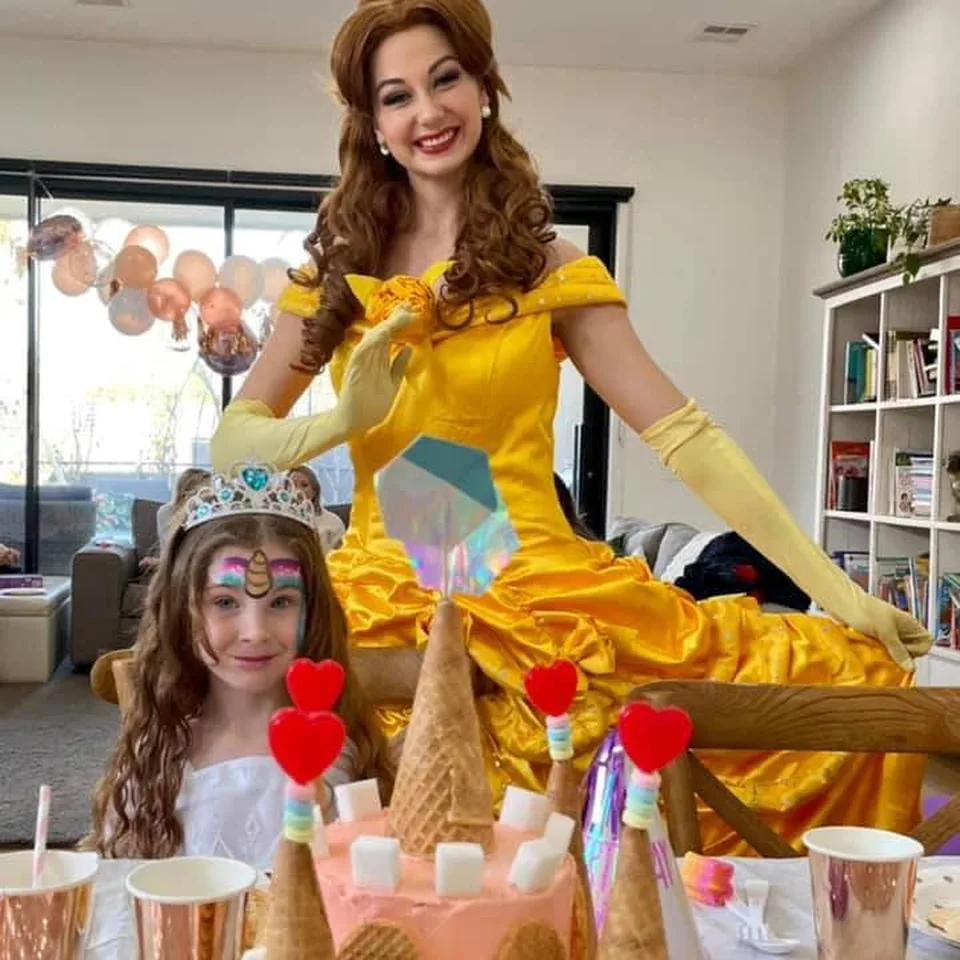 Princess Belle party entertainer in yellow Beauty and the Beast ballgown standing behind smiling girl in unicorn face paint and tiara beside a pink castle birthday cake at a Melbourne princess birthday party