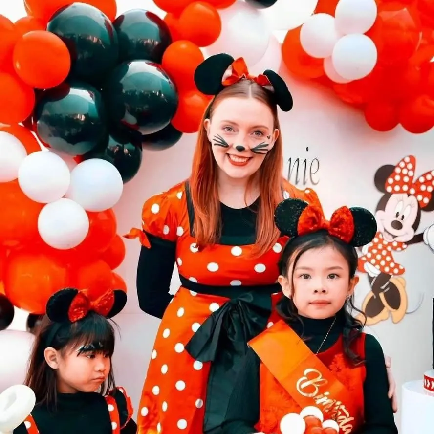 Minnie Mouse party entertainer with cat face paint and red polka dot dress posing with two young girls in Minnie ears at a red and black balloon arch in Melbourne