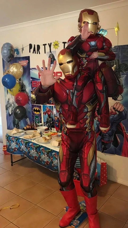 Iron Man party entertainer in full red and gold armoured suit holding young child in matching Iron Man costume on his shoulders in front of an Avengers party backdrop at a Melbourne superhero 4th birthday party