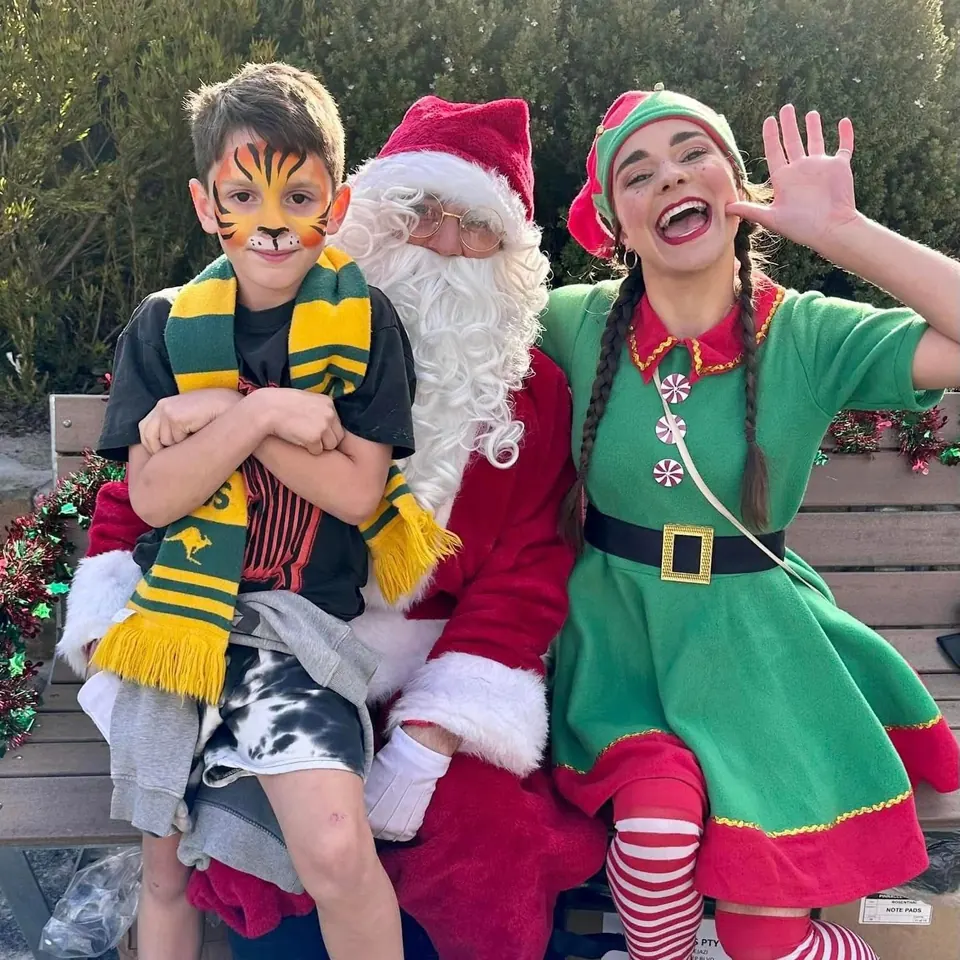 Santa and Christmas elf entertainers waving and laughing beside boy in tiger face paint and Wallabies scarf at a Melbourne Christmas community event