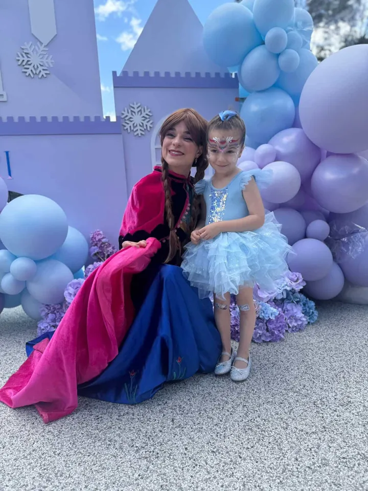 Frozen Anna party entertainer in pink cape and blue gown sitting with smiling young girl in a blue sequin and tulle dress and sparkly snowflake face paint in front of a purple Frozen castle backdrop and pastel balloon garland at a Melbourne Frozen birthday party