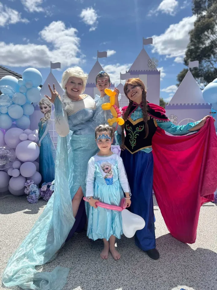 Frozen Elsa and Anna sister party entertainers in full Disney costumes posing with young girls in Elsa dresses and snowflake face paint in front of a purple Frozen castle backdrop and blue balloon garland at a Melbourne Frozen themed birthday party