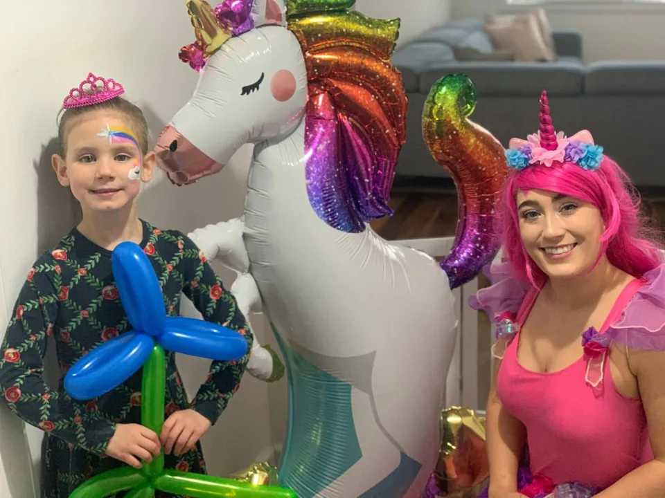 Unicorn fairy party entertainer with pink hair, rainbow horn and floral crown posing beside young girl in tiara holding a blue balloon flower at a Melbourne unicorn themed birthday party