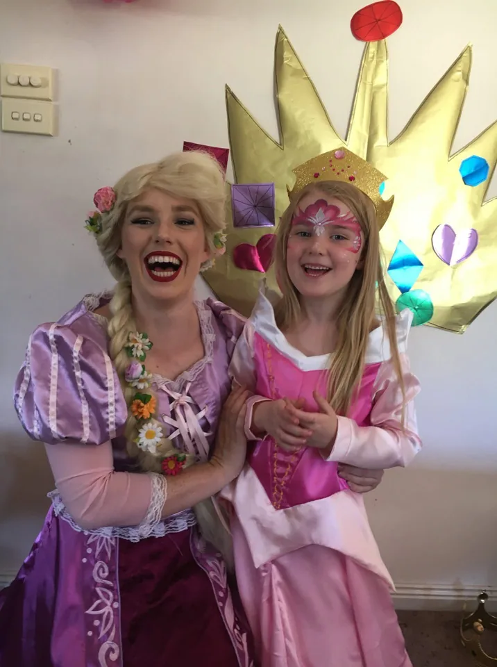 Princess Rapunzel party entertainer with long blonde braid and purple Tangled dress laughing with smiling girl in pink Aurora gown, gold crown and princess face paint at a Melbourne princess birthday party