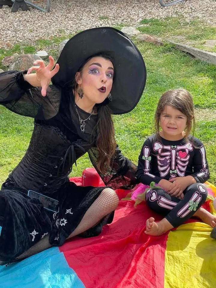 Witch character entertainer in black dress and hat with young girl in skeleton costume on a parachute mat at a Halloween-themed kids party in Melbourne