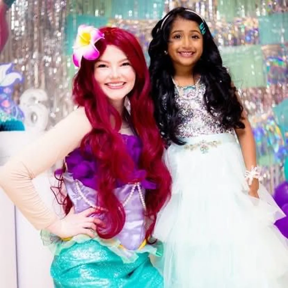 Little Mermaid Ariel party entertainer in purple and green sequin tail posing with smiling girl in white princess dress in front of mermaid scale backdrop at a Melbourne kids birthday party